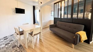 A modern living room with a sofa, a dining table with chairs, and a large window with blinds.