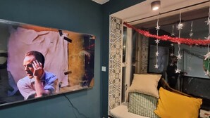 Een woonkamer met een grote flatscreen televisie aan de muur, een kamer met een patroon, een raam met decoratieve gordijnen en een bank met kussens.