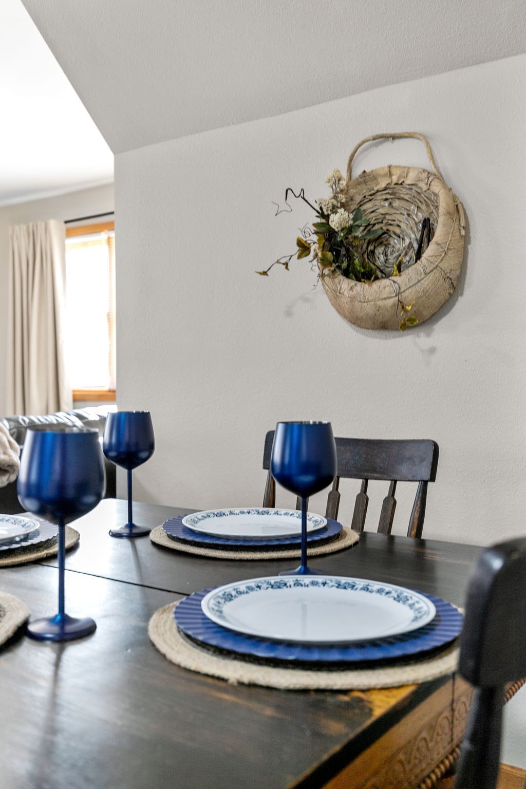 Une table à manger avec des verres à vin et des assiettes bleues, un panier en osier rempli de fleurs et une chaise en bois.
