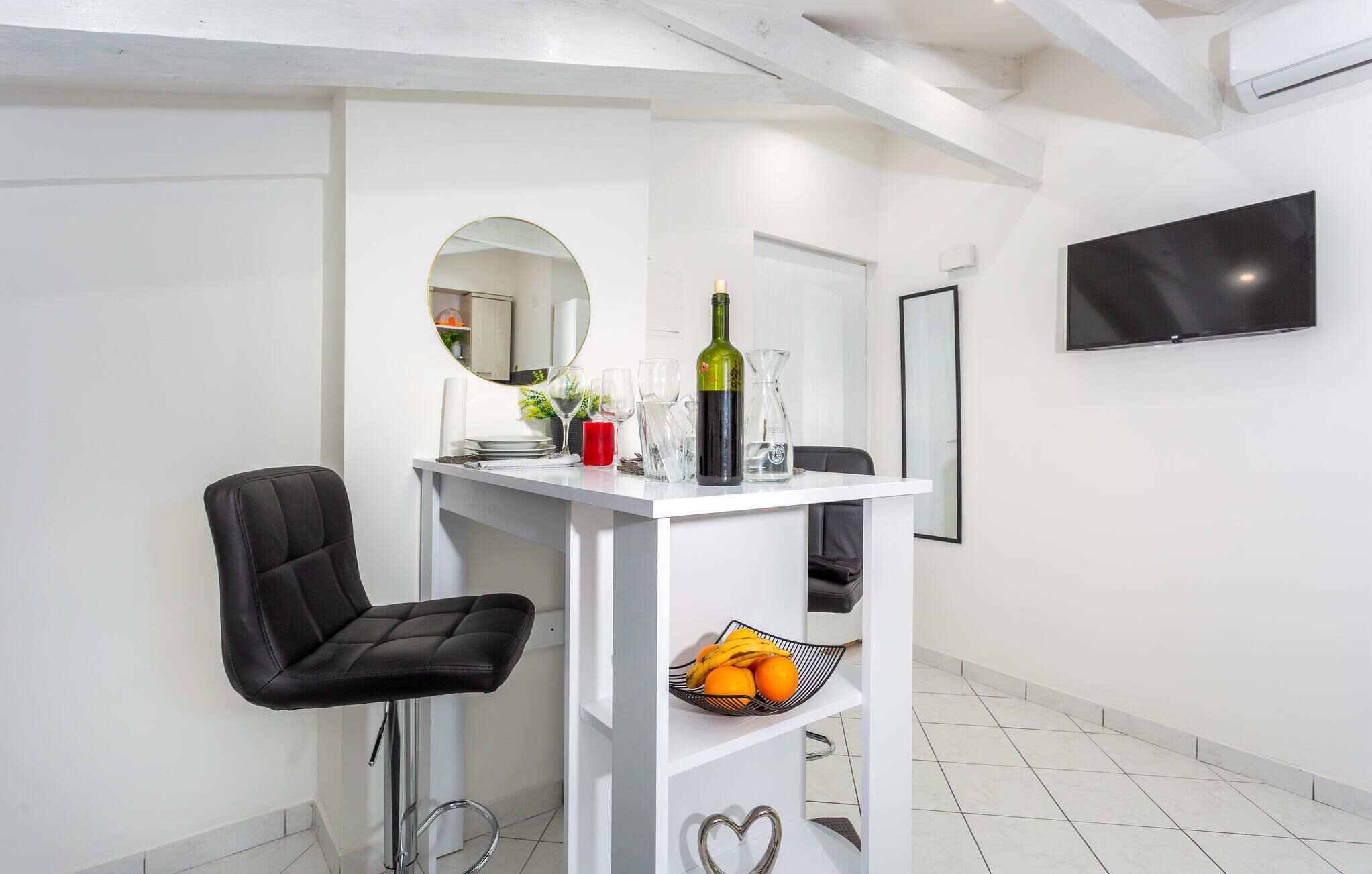 Une cuisine moderne avec des meubles blancs, un plan de travail blanc, une chaise noire, un téléviseur fixé au mur et un petit panier de fruits posé sur le plan de travail.