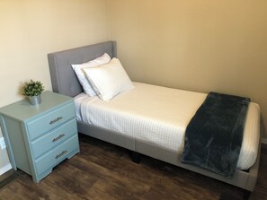 Una cama individual con cabecera gris, ropa de cama blanca y una manta oscura. Hay una mesita de noche azul claro con un cajón y una pequeña planta en maceta. El cuarto tiene piso de madera y una pared lisa.