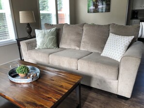 Un sofá color beige con varios cojines, una mesa de centro de madera con una bandeja y una planta, y una lámpara sobre una mesita auxiliar.
