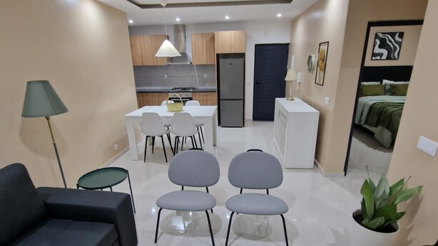 A modern living room with a sofa, a dining table with chairs, a kitchen area with cabinets and appliances, and a small balcony with a potted plant. - Zahma Apartments - Standard with 2 Bedrooms (Praia)