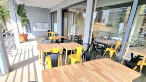 A modern indoor space with wooden tables and yellow chairs, large windows, and a potted plant. - Zahma Apartments - Standard with 2 Bedrooms (Praia)