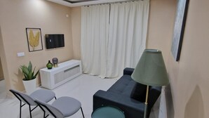 A modern living room with a sofa, two chairs, a small table, a floor lamp, a TV, and a potted plant. - Zahma Apartments - Standard with 2 Bedrooms (Praia)