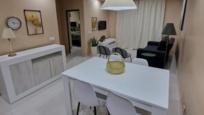 A modern dining area with a white table and chairs, a hanging light fixture, and a white console table with a clock and lamp. - Zahma Apartments - Standard with 2 Bedrooms (Praia)