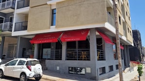 Un edificio de varios pisos con una cafetería en la planta baja, un coche blanco estacionado frente a él y un cielo despejado.