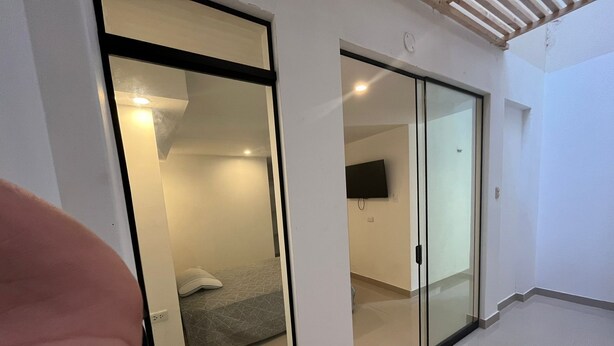 Une chambre d’hôtel moderne dotée d’une porte en verre donnant sur la salle de bain, d’un lit recouvert d’une couverture à motifs gris et d’un téléviseur à écran plat fixé au mur.