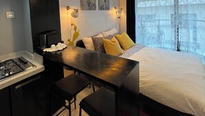 Quarto moderno com uma cama grande, uma mesa com duas cadeiras, uma área de cozinha e uma janela com vista para prédios.