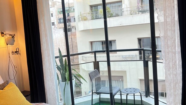Quarto com uma cama grande, uma planta em vaso e vista para uma varanda externa com cadeiras e uma mesa.