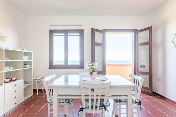 A dining area with a white table and chairs, a small potted plant, and a view of the sea through a window.