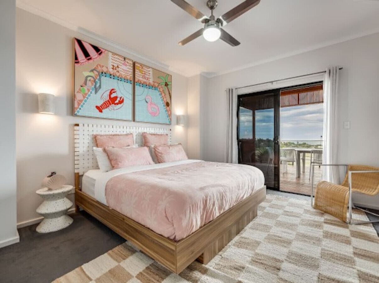 A bedroom with a bed, a ceiling fan, a large window with a view, a small round table, and a chair.