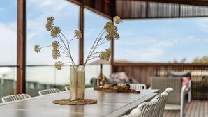 A dining table with a vase of dried flowers, surrounded by white chairs, on an outdoor deck with a view of the sky and a wooden fence in the background. - The Deck at Sellicks ~ Family friendly entertainer (Sellicks Beach)