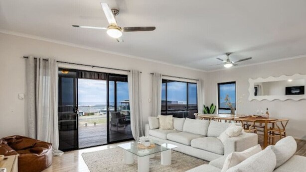 A spacious living room with a white sofa, a glass coffee table, and a dining area with a wooden table and chairs. - The Deck at Sellicks ~ Family friendly entertainer (Sellicks Beach)