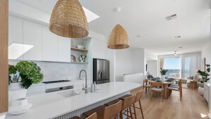 A modern kitchen with white cabinetry, a central island with bar stools, and a dining area with a view of the sea. - Horizons ~ Designer luxury stay with dream views (Aldinga Beach)