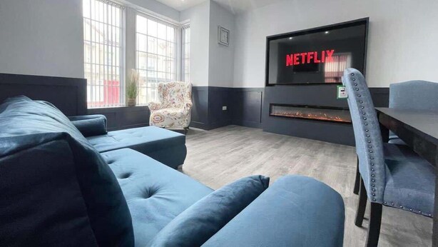 A modern living room with a large flat-screen TV displaying 'NETFLIX,' a blue sofa, a patterned armchair, and a dining table. - Serenity Wembley (Blackpool)