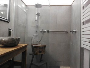 Une salle de bain avec un lavabo en pierre, une douche avec un pommeau de douche amovible, un porte-serviettes et un tabouret en bois muni d’un panier.