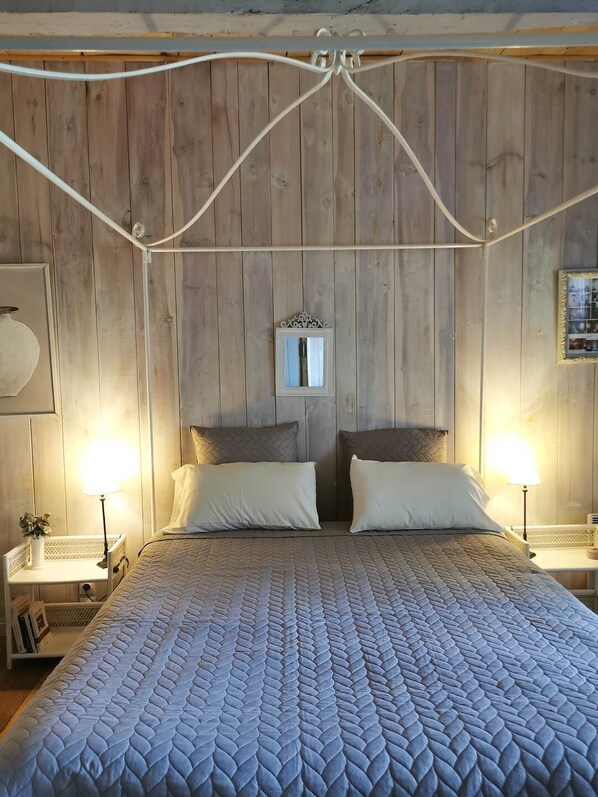 Une chambre à coucher avec une tête de lit en bois, deux tables de chevet avec des lampes, un grand lit recouvert d’une courtepointe, et un miroir au mur.