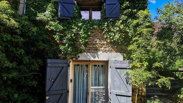 A rustic stone house with a wooden door and shutters, surrounded by lush greenery.