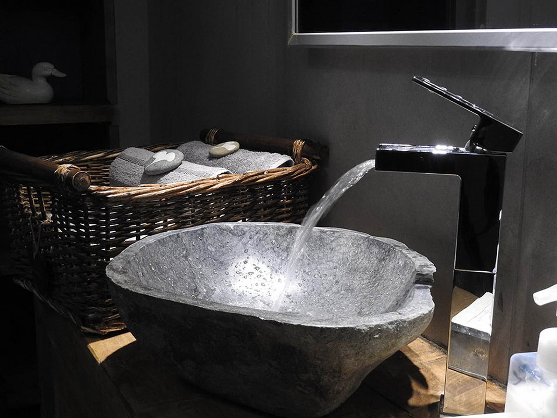 Un bassin en pierre dans lequel coule de l’eau, un panier en osier rempli de pierres et un robinet moderne.