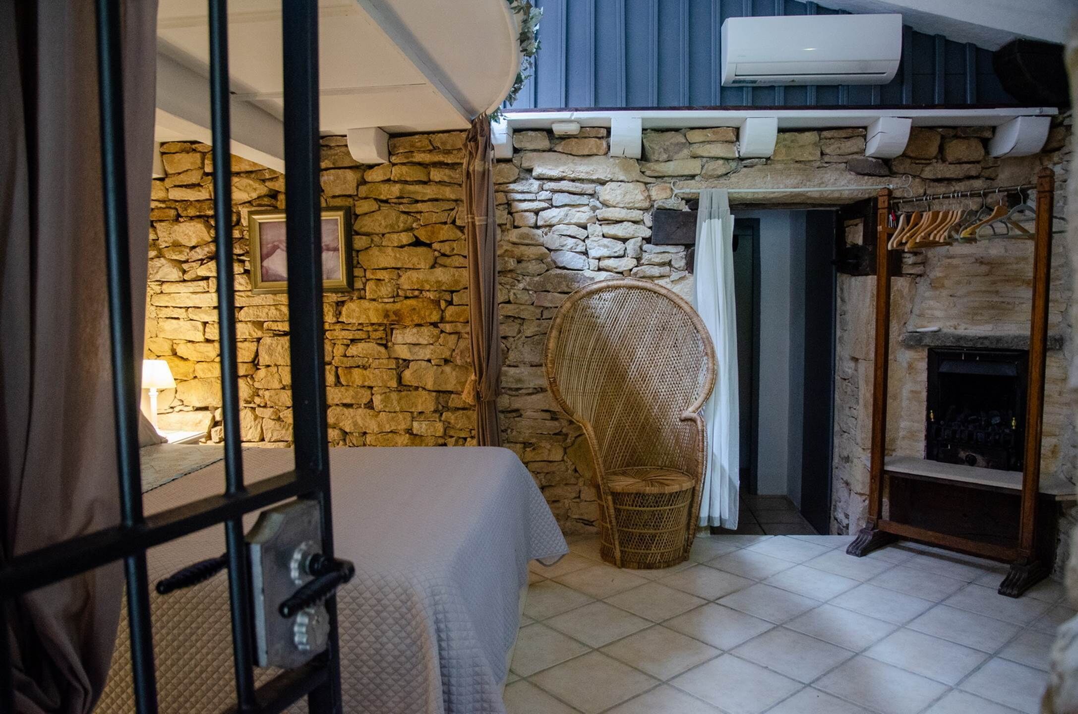 A cozy room with a stone wall, a wicker chair, a fireplace, and a bed with a white bedspread.