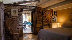 A bedroom with a stone wall, a wicker chair, a bed, and a view of an outdoor patio. - Guest House LA GRANGETTE (LUNAN)