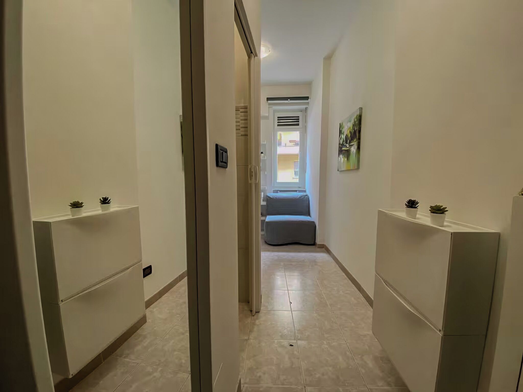 Ein schmaler Flur mit weißen Wänden und Fliesenboden, der zu einem Zimmer mit Bett und Fenster führt.