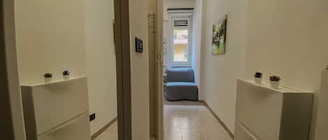 A narrow hallway with white walls and a tiled floor, leading to a room with a bed and a window.