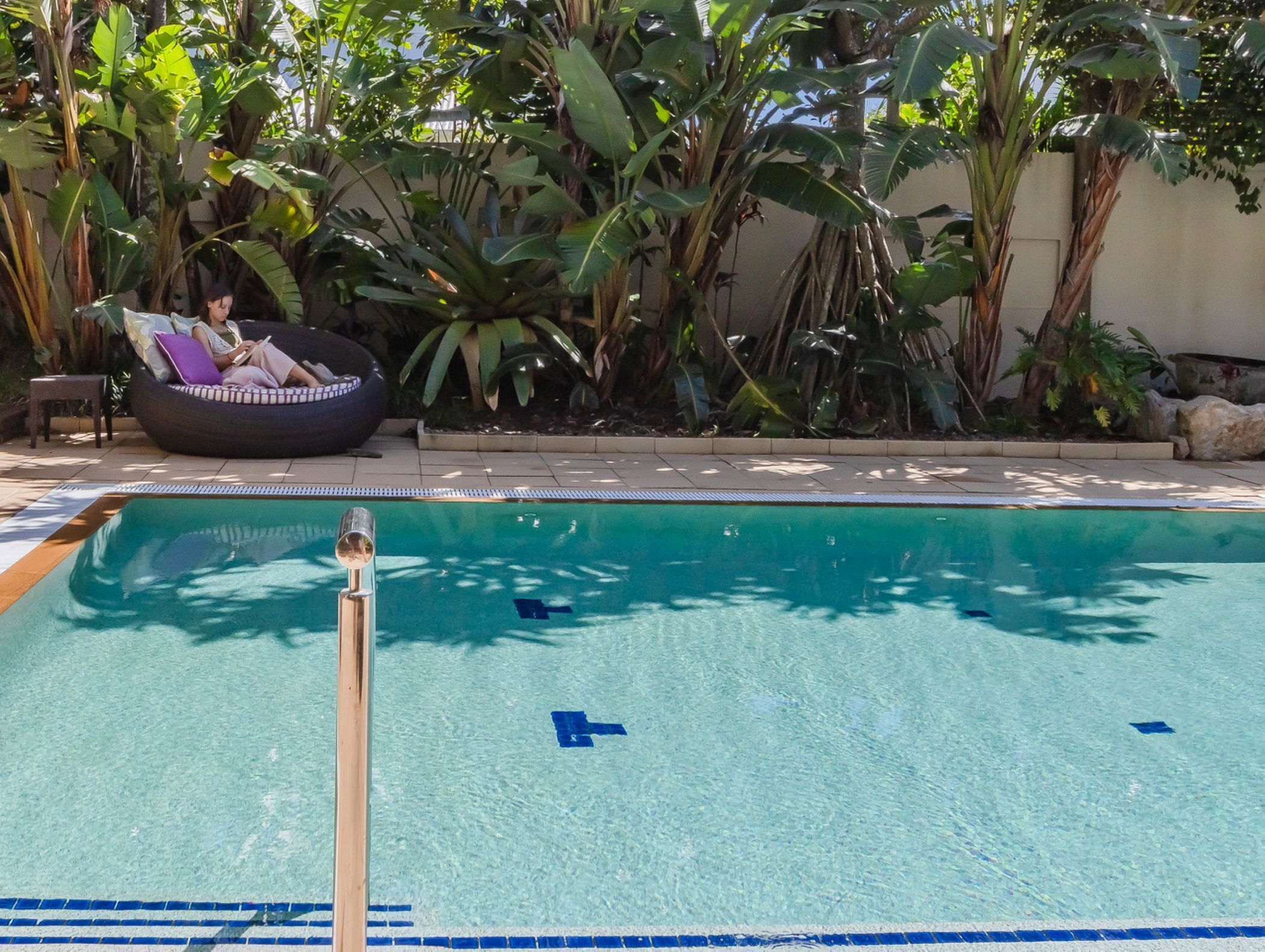 A swimming pool with clear blue water, surrounded by lush tropical plants and a lounge area with a bean bag chair.