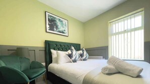 Una camera d'albergo con un letto, una poltrona verde, un quadro appeso e una finestra con persiane.
