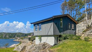 A wooden cabin with a large window, situated on a grassy hillside overlooking a body of water. - 2 bedroom amazing home in Mandal (Mandal)