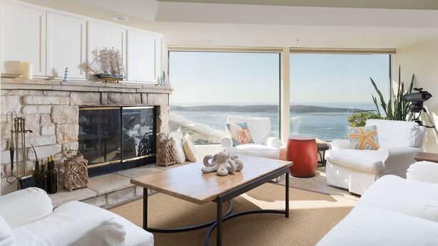 A modern living room with a stone fireplace, white furniture, and a large window overlooking the ocean.