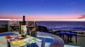 Une table avec deux verres à vin et une bouteille, donnant sur l’océan au coucher du soleil.