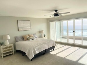 Une chambre à coucher comprenant un lit, des tables de chevet, un ventilateur de plafond et une grande fenêtre donnant sur l’océan.
