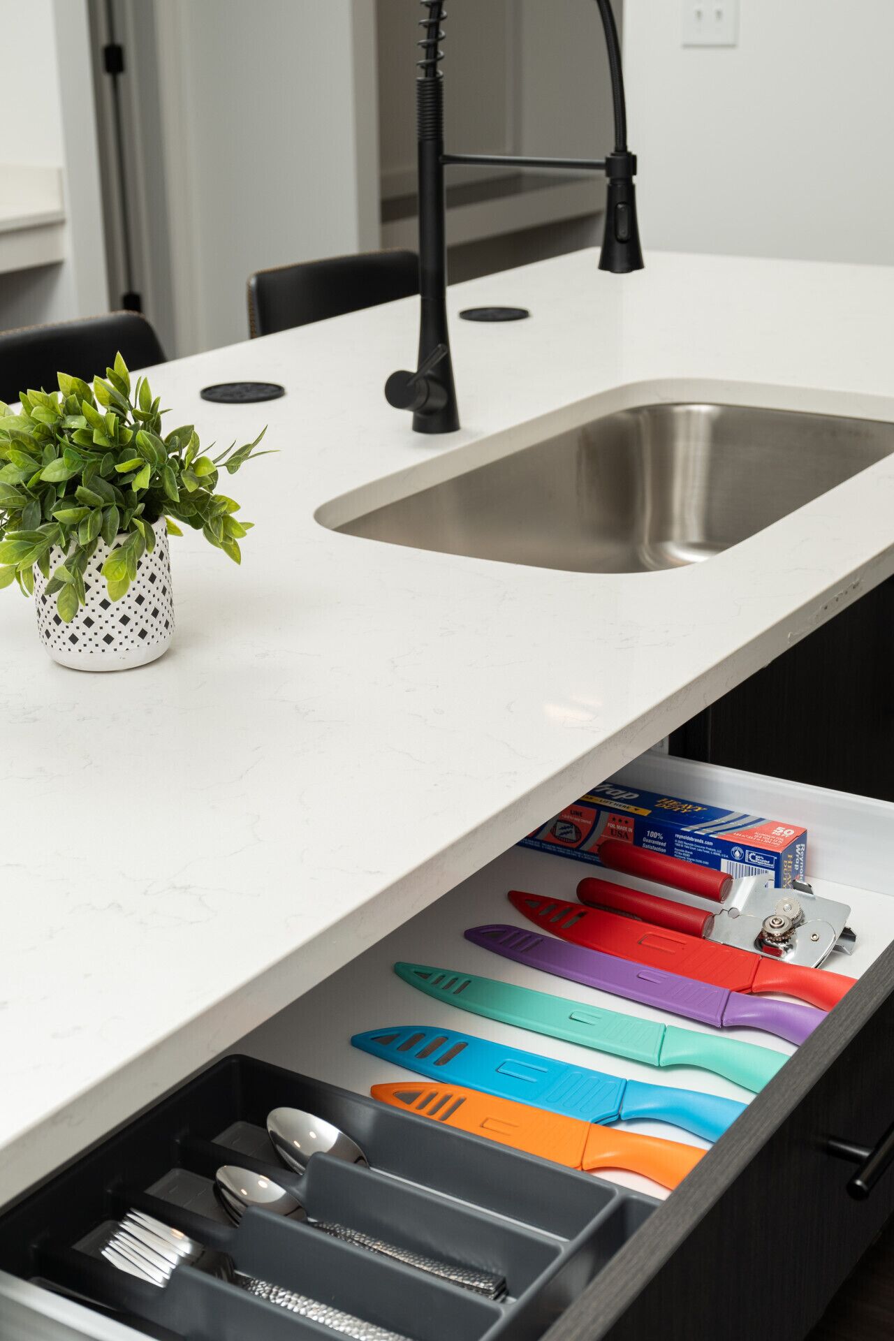 A kitchen with a white countertop, a stainless steel sink, and a drawer with colorful utensils.