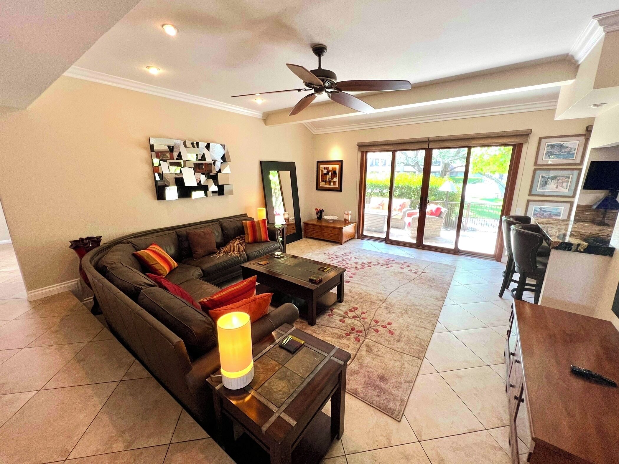 A spacious living room with a dark brown sofa, a coffee table, and a floor lamp. There is a ceiling fan, a large mirror, and a sliding glass door leading to an outdoor area.