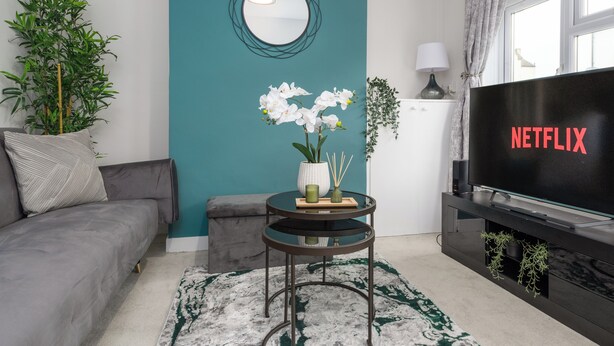 A modern living room with a grey sofa, a teal accent wall, a round mirror, a small round table with a vase of white flowers, a black TV with 'NETFLIX' on the screen, and a window with curtains. - Rochester Retreat - Charming 3 Bedroom House (Rochester)