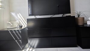 Ein modernes Wohnzimmer mit einem Flachbildfernseher, der an einer schwarzen Unterhaltungsecke befestigt ist. An der Wand befinden sich zwei kleine Regale mit Pflanzen, und ein dekorativer Vasen mit Federn steht auf dem Beistelltisch.
