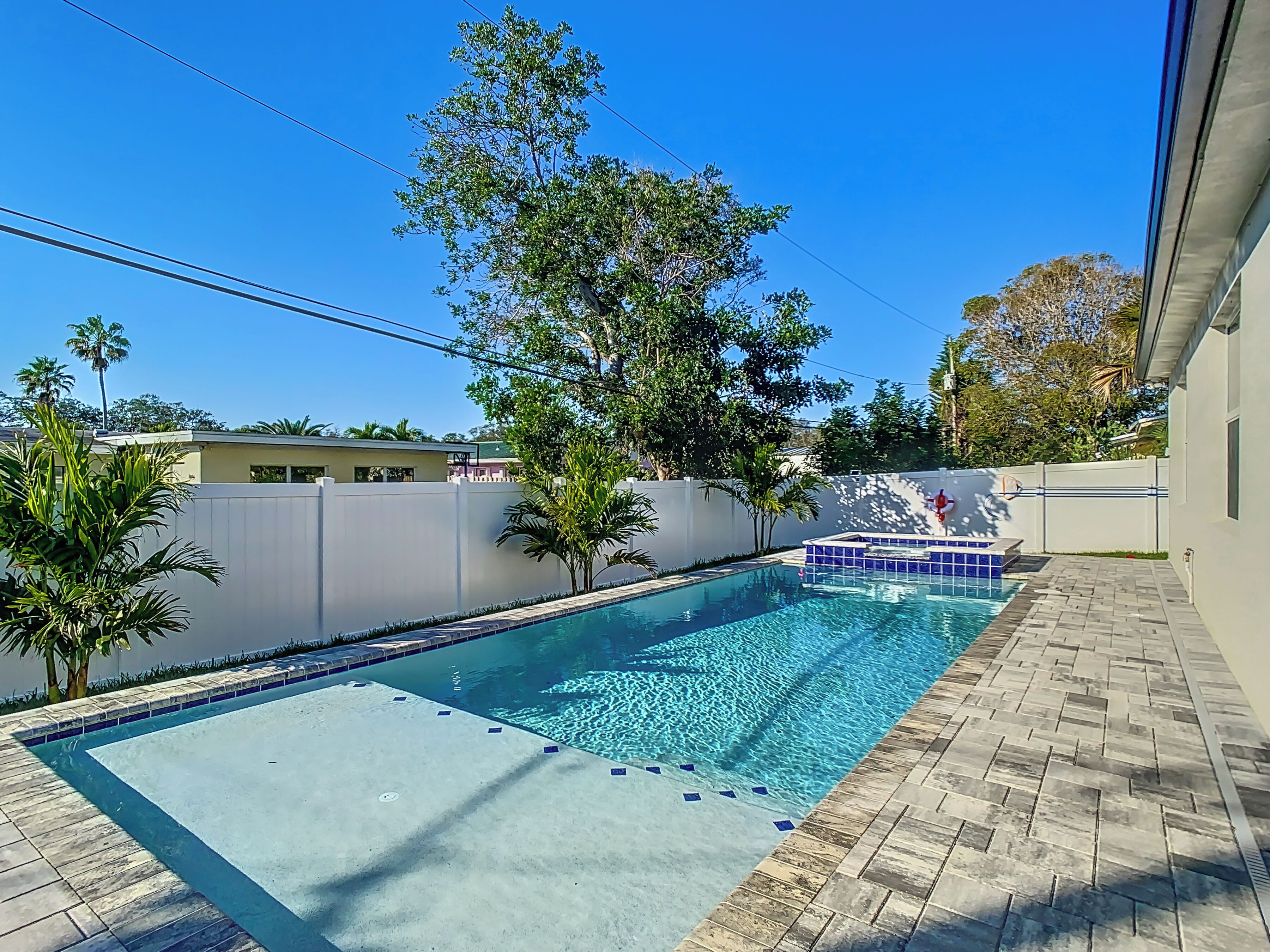 A rectangular swimming pool with clear blue water, surrounded by a white fence and trees.