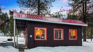 A small, dark wooden cabin with a red roof and two windows with orange frames, surrounded by snow. - Private Hot Spring, Perfect for Couples, No Shared Spaces, 1 Hour North of Boise (Garden Valley)