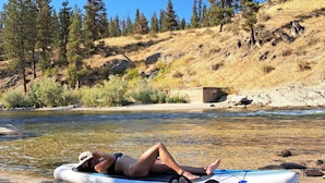 A person lying on a lounge chair by a river, with a paddleboard nearby. - Private Hot Spring, Perfect for Couples, No Shared Spaces, 1 Hour North of Boise (Garden Valley)