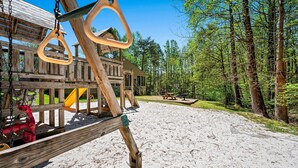 A wooden play structure with swings and a slide, surrounded by a forested area. - 🔥 Mountain Escape | Fireplace, Sky Deck & Year-Round Resort Perks! (Gatlinburg)