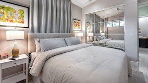 A modern hotel room with a large bed, a nightstand with a lamp, a mirror, and a framed picture on the wall.
