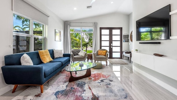 A modern living room with a blue sofa, two armchairs, a glass coffee table, and a TV mounted on the wall.