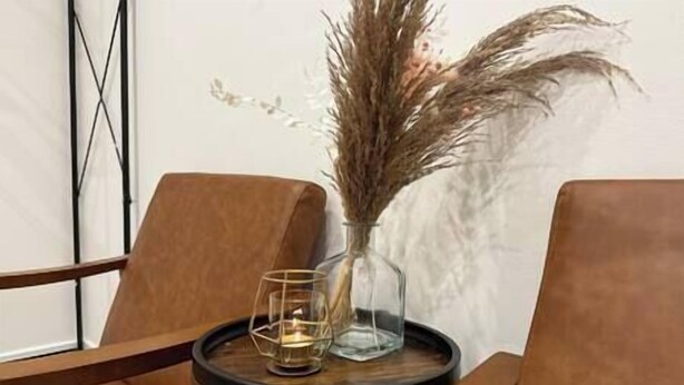 Une chaise en bois, une table en verre, un vase en verre transparent contenant des herbes de la pampa séchées et un chandelier en verre.