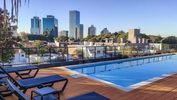 Piscina en la azotea con cubierta de madera y sillones de descanso, con vista a un horizonte urbano con edificios modernos.