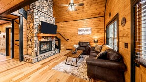 A cozy living room with a stone fireplace, leather sofa, and a ceiling fan. - New! Woodridge Lodge! 3 min to PF Pkwy! Pool,Pool Table,Hot Tub,Shuffboard,Poker (Pigeon Forge)