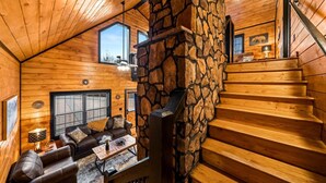 A log cabin interior with a stone fireplace, wooden staircase, and a cozy living area with a sofa and coffee table. - New! Woodridge Lodge! 3 min to PF Pkwy! Pool,Pool Table,Hot Tub,Shuffboard,Poker (Pigeon Forge)
