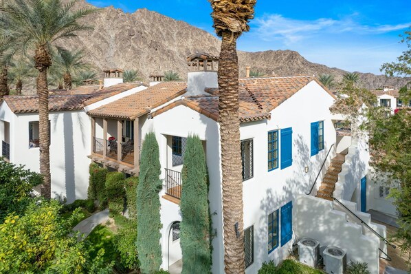 A white building with blue shutters, surrounded by palm trees and cypress trees, with a mountainous backdrop.