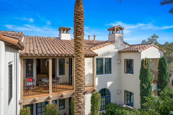 Una casa de dos pisos con techo de tejas, un balcón con mobiliario exterior y una palmera alta al frente.